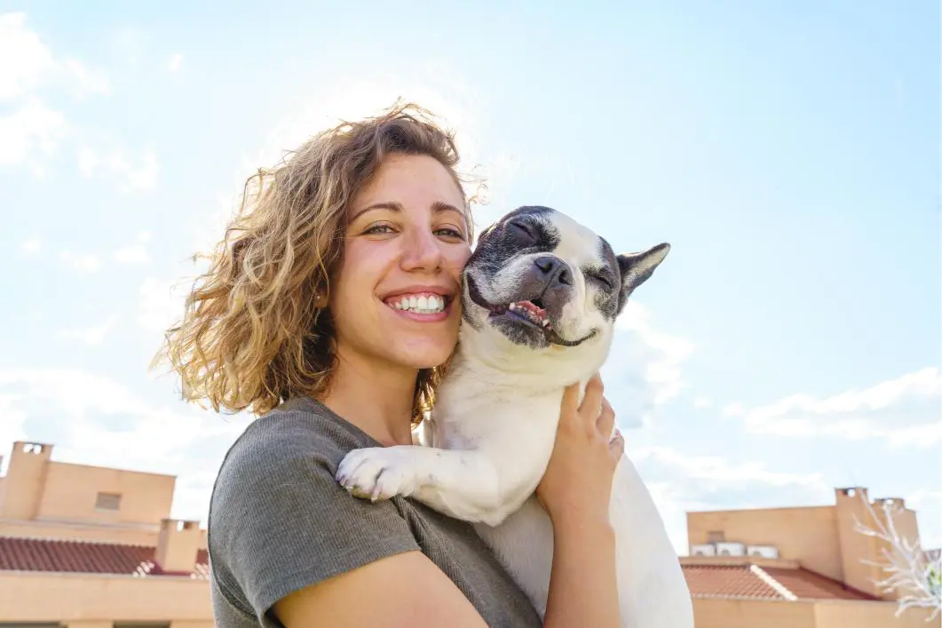 GettyImages-1334498420 A person smiling and holding a small dog outdoors on a sunny day with blue skies and some clouds in the background.