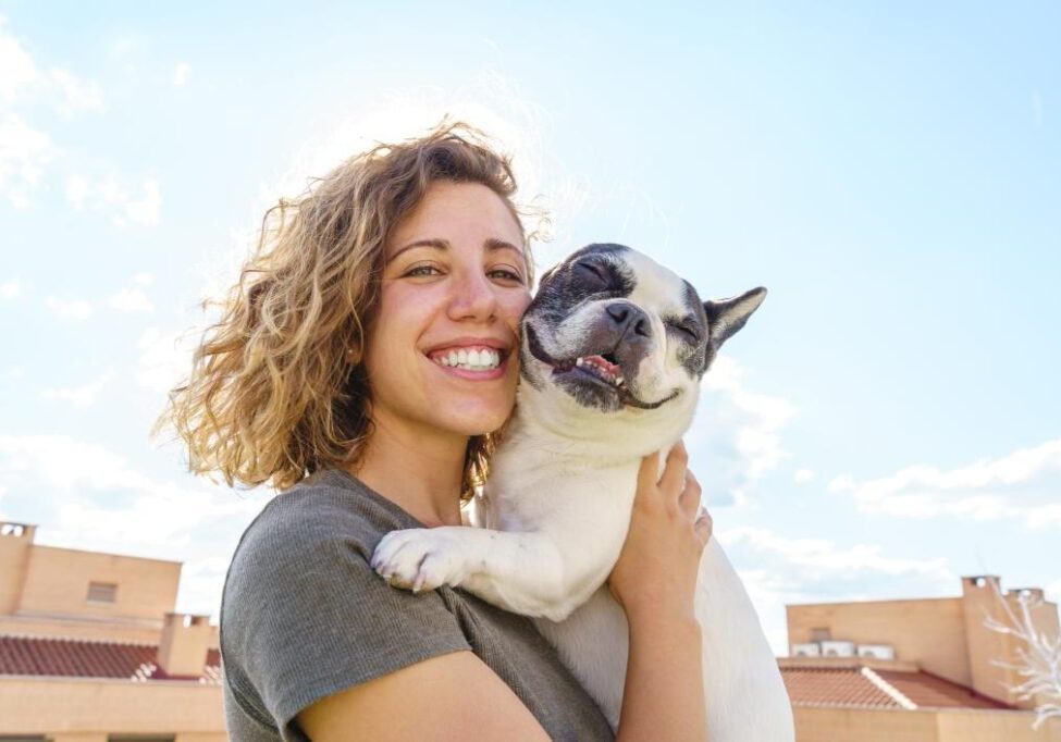 A person smiling and holding a small dog outdoors on a sunny day with blue skies and some clouds in the background.